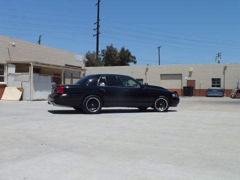 2003 Ford Crown Victoria - Side profile of a lowered black Ford Crown Victoria with dark wheels parked before low industrial buildings.