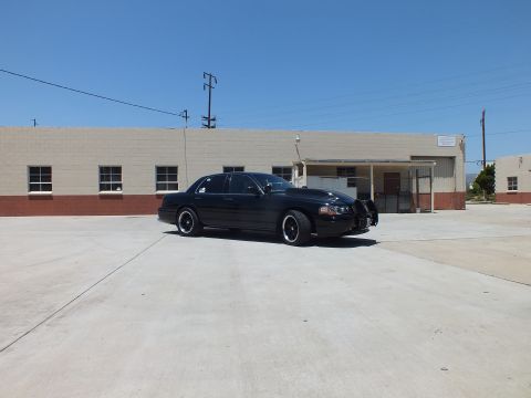 2003 Ford Crown Victoria - black sedan angled across a wide concrete yard in front of a tan industrial building under blue sky.