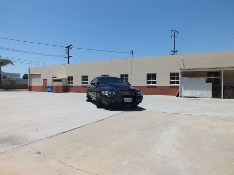 2003 Ford Crown Victoria - dark sedan with a front push bar sits at distance in an expansive concrete lot beside a tan building.