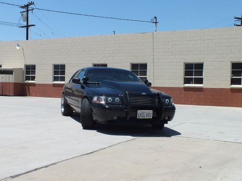 2003 Ford Crown Victoria - black Ford Crown Victoria with a push bar carries a California plate reading EVIL VIC in a brick-trimmed lot.
