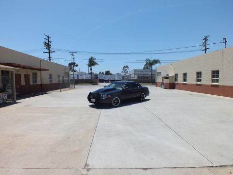 2003 Ford Crown Victoria - black sedan with a front push bar sits centered in a wide empty concrete lot ringed by industrial buildings.
