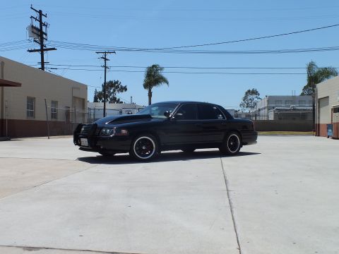 2003 Ford Crown Victoria - Side-front view of a lowered black Ford Crown Victoria with a push bar and dark wheels in a sunny lot.
