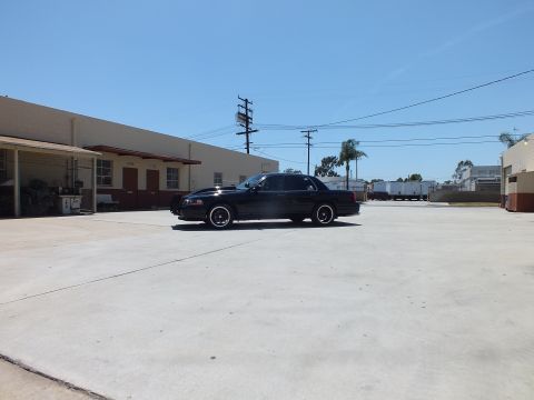 2003 Ford Crown Victoria - black sedan sits in profile far across a wide concrete yard with trucks and palm trees in the background.