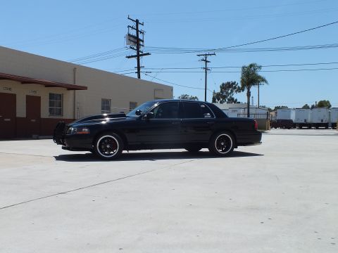 2003 Ford Crown Victoria - Full side profile of a black Ford Crown Victoria with a push bar parked before a tan industrial building.