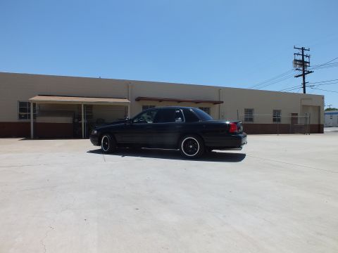 2003 Ford Crown Victoria - black sedan with a trunk spoiler angles beside a tan warehouse with a carport in a concrete lot.