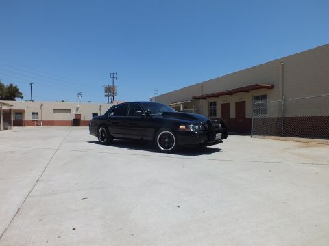 2003 Ford Crown Victoria - black Ford Crown Victoria with a push bar sits in a concrete lot beside a chain-link fence and tan buildings.