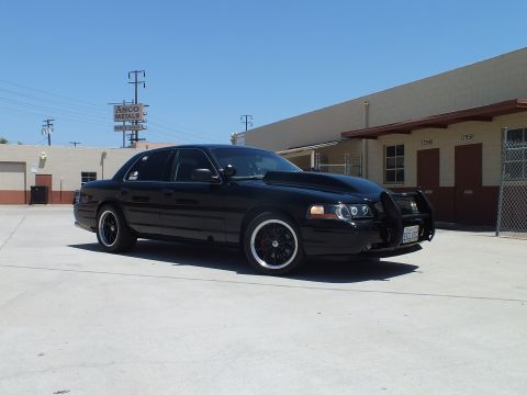 2003 Ford Crown Victoria - black Ford Crown Victoria with a push bar parks before tan buildings and an ASCO Metals sign.