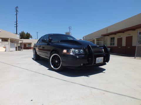 2003 Ford Crown Victoria - black Ford Crown Victoria with a push bar and EVIL VIC plate angles across a sunny concrete lot.
