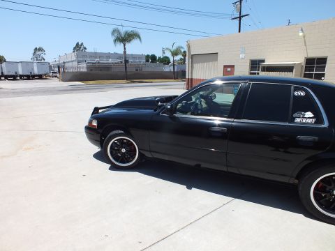 2003 Ford Crown Victoria - Side view of a black sedan with rear-window decals and dark wheels parked beside a tan industrial building.