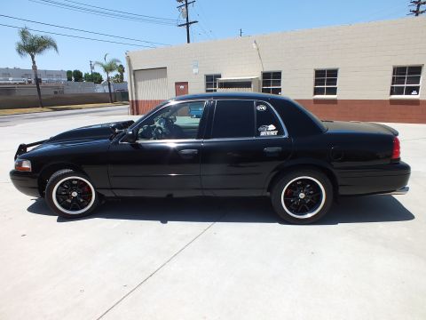 2003 Ford Crown Victoria - Complete side profile of a black Ford Crown Victoria with dark wheels parked before a tan industrial building.