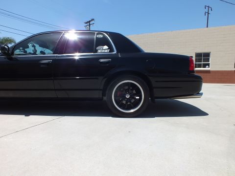 2003 Ford Crown Victoria - Low rear-quarter view of a black sedan with window decals and a black alloy wheel, bright sun flare overhead.