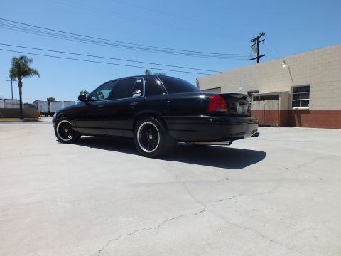 2003 Ford Crown Victoria - blacked-out full-size sedan sits in an empty concrete lot beside warehouse buildings under a clear blue sky.