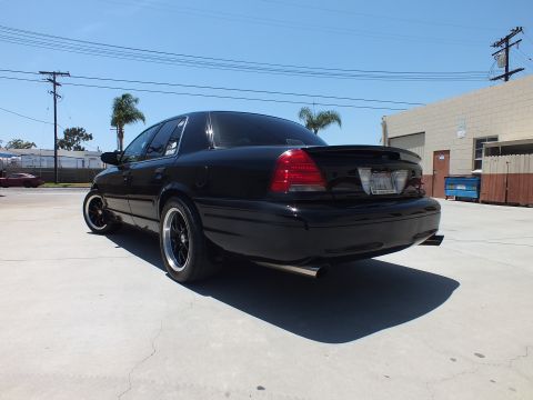 2003 Ford Crown Victoria - glossy black sedan with dark wheels and a chrome dual exhaust tip is parked rear-quarter in front of warehouses.