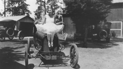 1909 Ford Model T - A woman dressed as a ballerina poses on the hood of a 1909 Ford Model T, with another car visible in the background.
