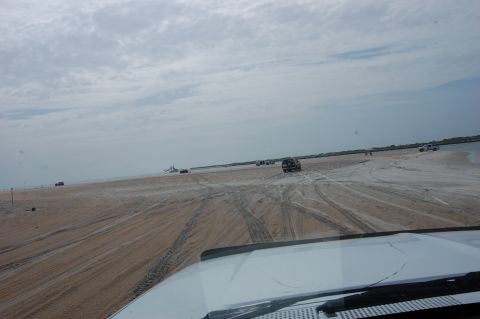 1994 Ford Bronco - Multiple vehicles drive on a sandy beach under a cloudy sky, with the foreground showing the interior of a white SUV.