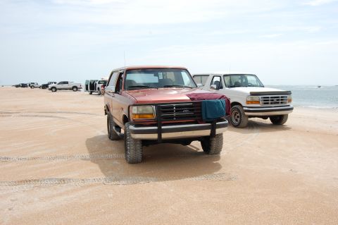 1994 Ford Bronco - Two Ford Broncos parked on a sandy beach near the ocean.