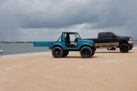 1994 Ford Bronco - Two modified vehicles are parked on a beach with a cloudy sky overhead.
