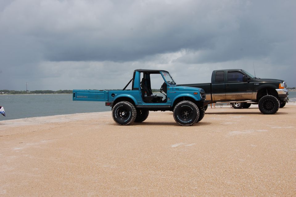 Blue Suzuki Samurai flatbed and black pickup truck on a sandy beach near the water