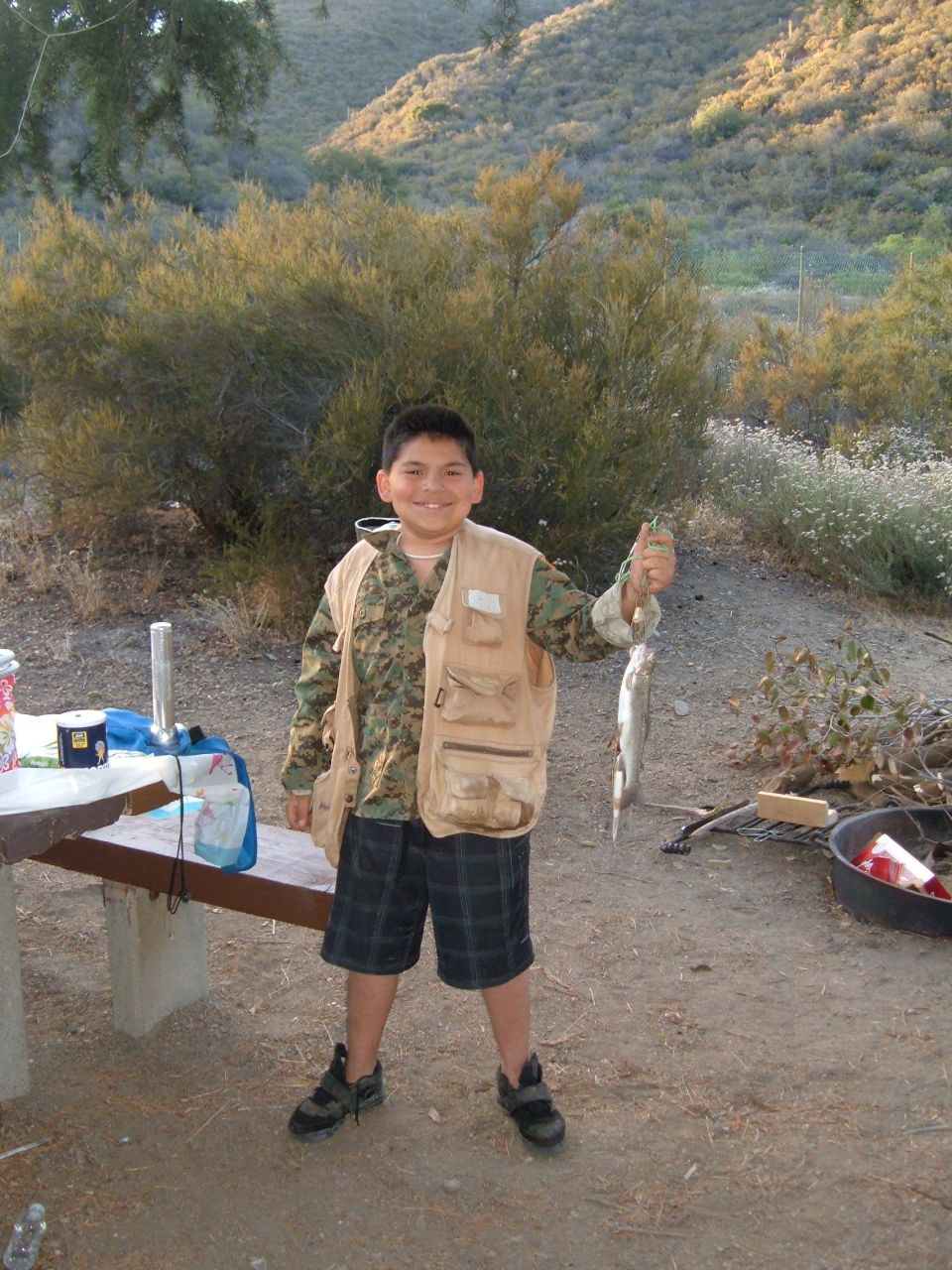 Kid in fishing vest holds up a small fish he caught, standing in a dry, hilly area with bushes and a picnic table nearby