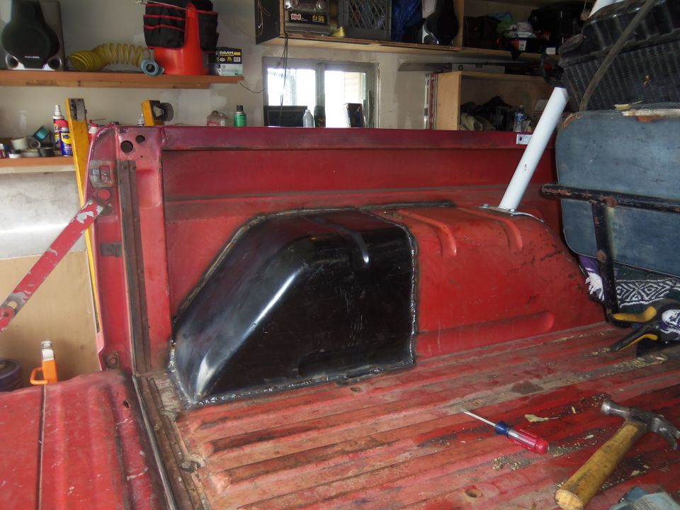 Truck bed interior showing a black fuel tank cover installed over the wheel well. Red paint is visible on the bed floor and sides