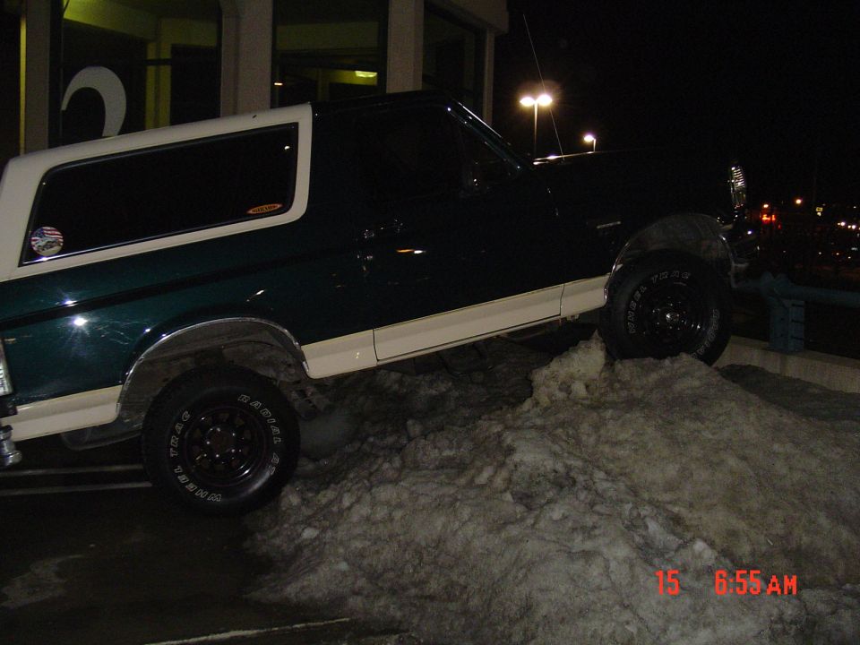 Green and white full-size Bronco climbing a dirty snow pile at night in a lit parking lot