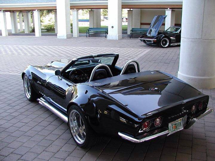 Black convertible Corvette from rear-right angle, chrome side pipes, aftermarket wheels, another Corvette with hood up in background