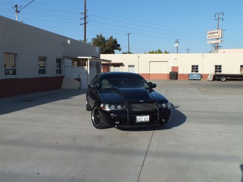 2003 Ford Crown Victoria - black sedan with a murdered-out front grille and dark wheels faces the camera in a wide concrete lot by warehouses.