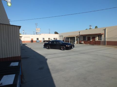 2003 Ford Crown Victoria - small black sedan sits across a broad sunlit industrial lot framed by warehouse buildings and power lines.