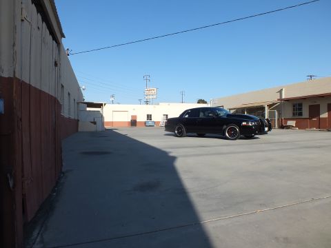 2003 Ford Crown Victoria - black sedan sits along a warehouse wall in a wide concrete lot with overhead wires and a clear sky.
