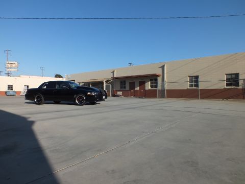 2003 Ford Crown Victoria - black full-size sedan is parked at a front-quarter angle in a sunlit industrial yard beside long warehouse buildings.