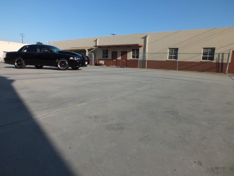 2003 Ford Crown Victoria - black sedan with dark wheels sits at a front-quarter angle in a wide concrete lot beside a tan warehouse.