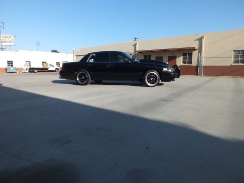 2003 Ford Crown Victoria - black full-size sedan shows its full driver-side profile, lowered on black wheels, in a sunlit industrial lot.