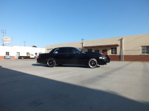 2003 Ford Crown Victoria - black full-size sedan is shown in clean side profile on black wheels across an open industrial concrete lot.