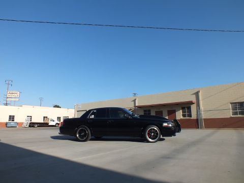 2003 Ford Crown Victoria - black sedan in low side profile sits on dark wheels beneath a deep blue sky in front of warehouse buildings.