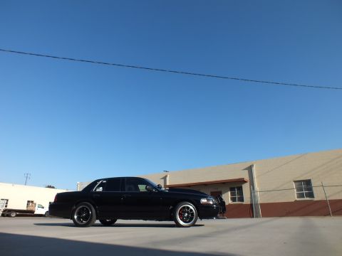 2003 Ford Crown Victoria - low-angle front-quarter shot of a black sedan emphasizes its dark wheels and red brake calipers under a blue sky.
