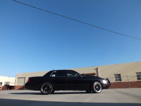2003 Ford Crown Victoria - black sedan with one headlight lit sits low on dark wheels in a concrete lot under a vivid blue sky.