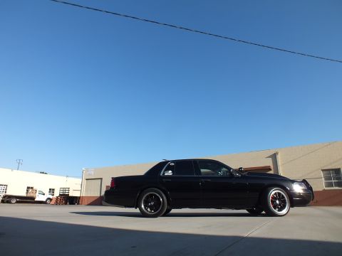 2003 Ford Crown Victoria - black full-size sedan with headlights on poses at a low front-quarter angle across a wide industrial concrete lot.
