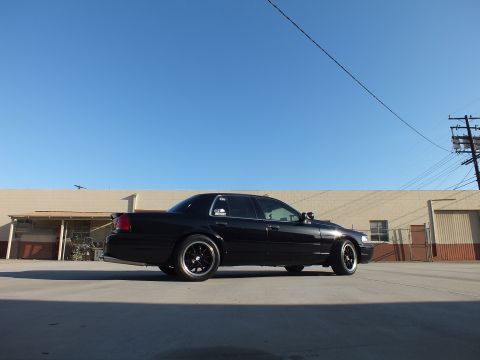 2003 Ford Crown Victoria - low-angle rear-quarter shot shows a black sedan on dark wheels against a tan warehouse and clear blue sky.