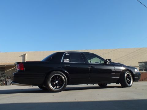 2003 Ford Crown Victoria - black full-size sedan shows its passenger-side rear-quarter, with window decals and black wheels, in an industrial lot.
