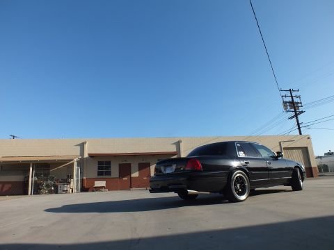 2003 Ford Crown Victoria - black sedan in low rear-quarter view sits beside a long warehouse with overhead power lines and a blue sky.
