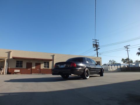 2003 Ford Crown Victoria - black full-size sedan is parked in rear three-quarter view with glowing taillights beside warehouse buildings.