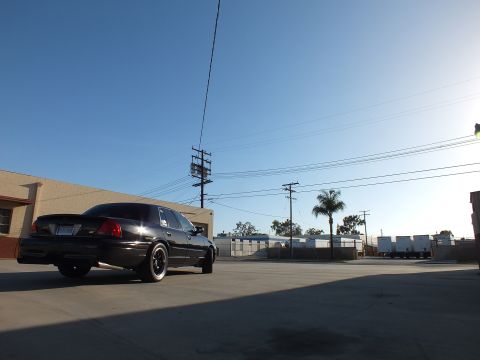 2003 Ford Crown Victoria - black sedan sits in rear-quarter view backlit by a low sun across a wide industrial lot with power poles.