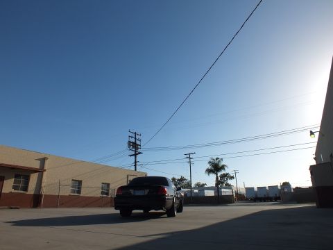 2003 Ford Crown Victoria - black sedan is parked rear-on across a sunlit industrial lot framed by warehouses, power poles, and a palm tree.