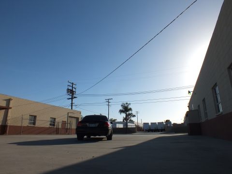 2003 Ford Crown Victoria - small black sedan sits far across a broad concrete industrial lot with warehouses, power lines, and bright sun.