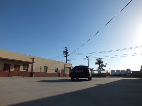 2003 Ford Crown Victoria - black full-size sedan sits alone in a sunlit industrial parking lot beside a tan warehouse with power lines overhead.