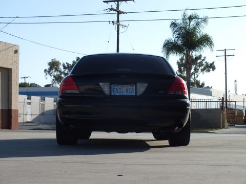 2003 Ford Crown Victoria - Rear view of a black sedan showing taillights and a California license plate reading EVIL VIC, parked near a palm tree and warehouses.