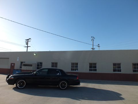 2003 Ford Crown Victoria - Side profile of a lowered black full-size sedan with dark wheels parked along a tan warehouse wall.