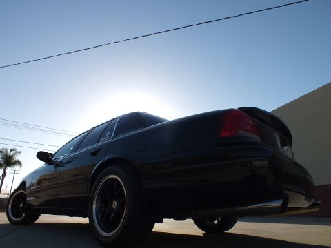 2003 Ford Crown Victoria - Dramatic low rear-quarter shot of a black sedan with black-and-polished wheels and a small rear lip spoiler, backlit by sun.
