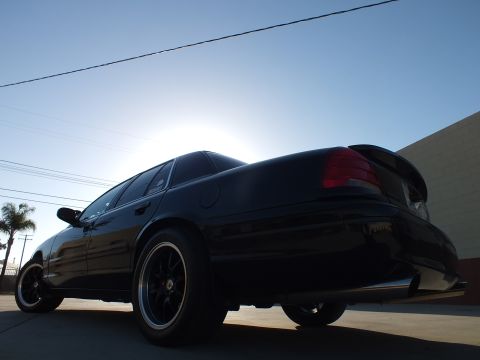 2003 Ford Crown Victoria - Low-angle rear-quarter view of a black sedan with glowing red taillights and dark wheels against a bright sky.