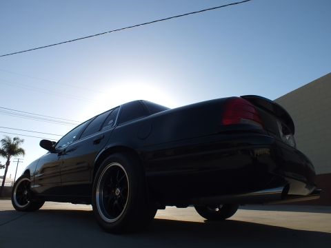 2003 Ford Crown Victoria - black sedan photographed from a low rear angle, showing its rear lip spoiler, dark wheels and tinted windows in strong sun.